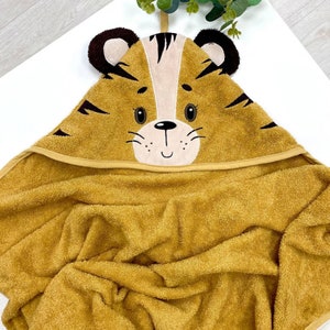 May include: A mustard yellow hooded towel for babies, featuring a tiger face design on the hood. The tiger's face has brown and tan details, black stripes, and a smiling mouth. The towel is made from a soft, absorbent fabric, perfect for after bath time.