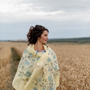 May include: A woman wearing a yellow floral shawl with blue accents stands in a field of wheat. The shawl is draped over her shoulders and she is looking off to the side.
