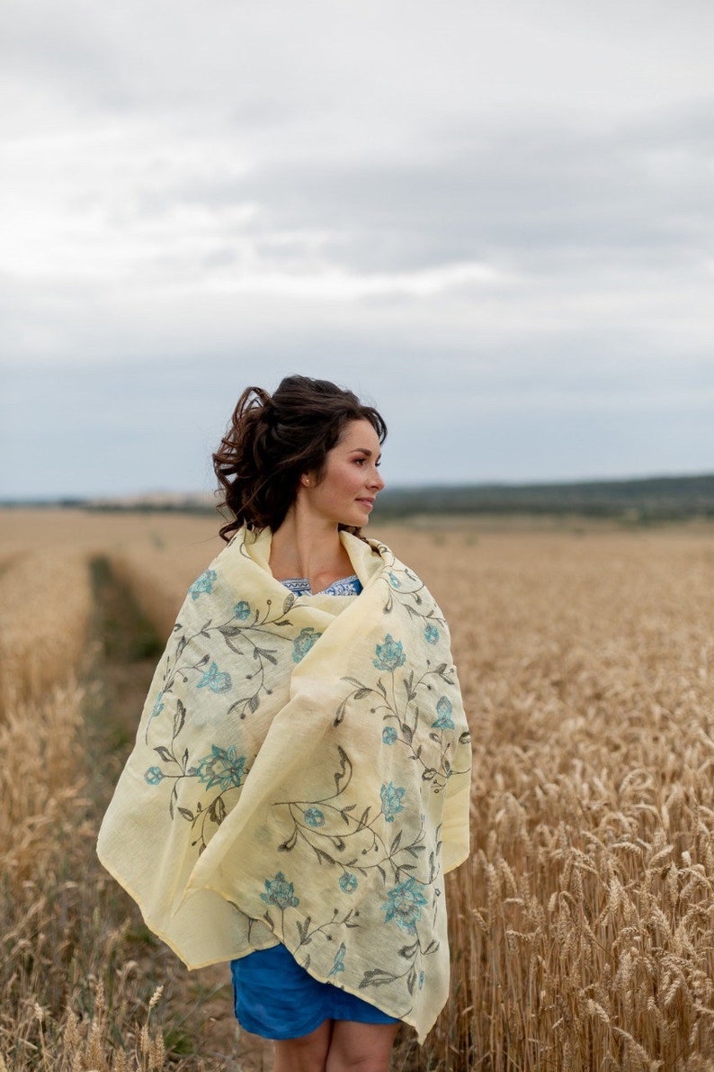 May include: A woman wearing a yellow floral shawl with blue accents stands in a field of wheat. The shawl is draped over her shoulders and she is looking off to the side.
