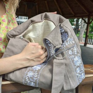 Peut inclure: Un sac fourre-tout en toile beige clair avec une fermeture &eacute;clair et une longue bandouli&egrave;re. Le sac est doubl&eacute; de blanc et d&eacute;cor&eacute; de fleurs bleues brod&eacute;es.