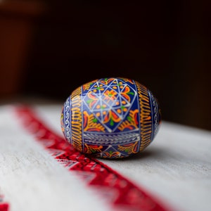 K&ouml;nnte beinhalten: Ein verziertes Ei mit blauem Hintergrund und komplizierten Mustern in Orange, Gelb und Gr&uuml;n. Das Ei liegt auf einem wei&szlig;en Tuch mit einem rot bestickten Rand, das traditionelle Handwerkskunst zeigt.