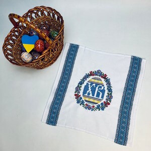 May include: A white Easter towel with blue embroidered borders and a central design of a decorated egg with the letters "XP" in blue. A wicker basket filled with decorated Easter eggs sits nearby.