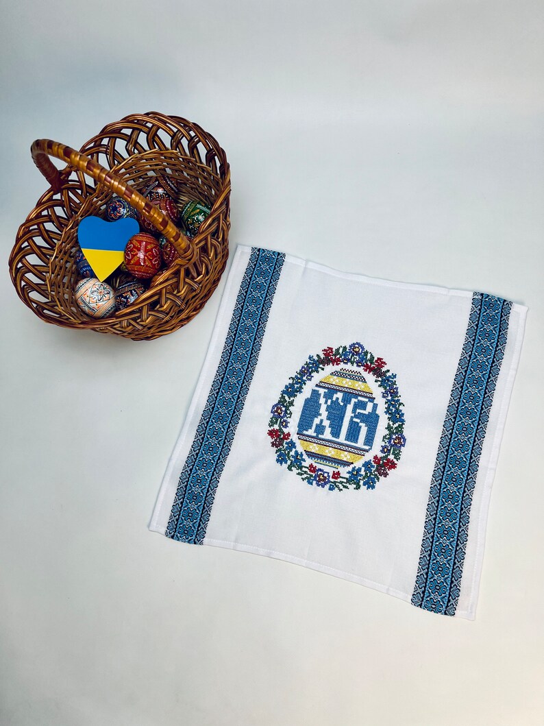 May include: A white Easter towel with blue embroidered borders and a central design of a decorated egg with the letters "XP" in blue. A wicker basket filled with decorated Easter eggs sits nearby.
