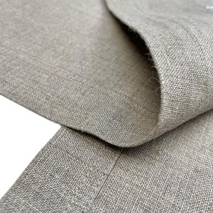 May include: Close-up of a section of tan linen fabric, showing the texture and weave. The fabric is folded over, revealing the inside and the edge of the material. The neutral color and natural fibers suggest a garment or home textile.