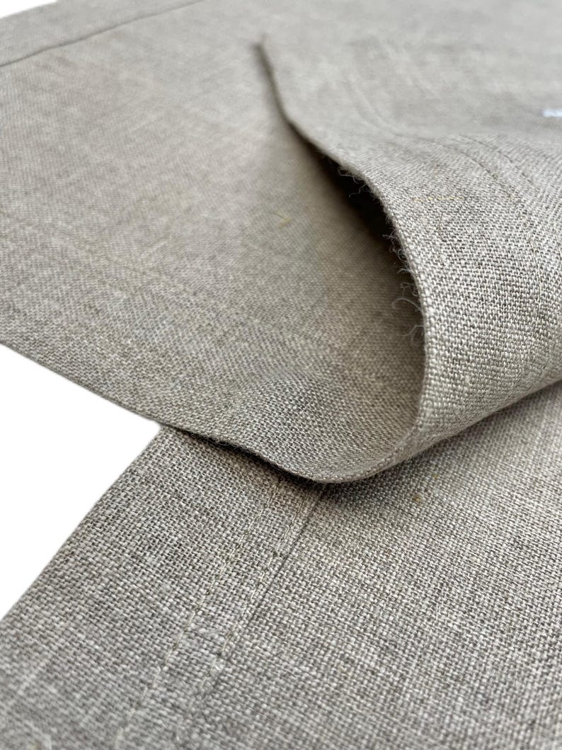 May include: Close-up of a section of tan linen fabric, showing the texture and weave. The fabric is folded over, revealing the inside and the edge of the material. The neutral color and natural fibers suggest a garment or home textile.