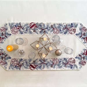 Puede incluir: Una mesa navide&ntilde;a festiva con un mantel blanco, un camino de mesa decorativo con adornos y manteles individuales a juego. Cubiertos dorados, platos blancos y vasos transparentes completan la decoraci&oacute;n navide&ntilde;a. Un candelabro geom&eacute;trico y velas encendidas a&ntilde;aden calidez.