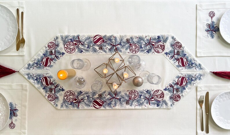 Puede incluir: Una mesa navide&ntilde;a festiva con un mantel blanco, un camino de mesa decorativo con adornos y manteles individuales a juego. Cubiertos dorados, platos blancos y vasos transparentes completan la decoraci&oacute;n navide&ntilde;a. Un candelabro geom&eacute;trico y velas encendidas a&ntilde;aden calidez.