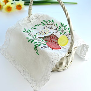 May include: White wicker basket with a handle, lined with a white cloth featuring an embroidered Easter design. The embroidery includes a red egg, yellow egg, and green leaves. The text "Style 1" is visible.