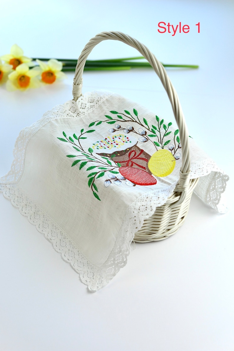 May include: White wicker basket with a handle, lined with a white cloth featuring an embroidered Easter design. The embroidery includes a red egg, yellow egg, and green leaves. The text "Style 1" is visible.