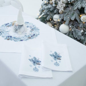 K&ouml;nnte beinhalten: Ein wei&szlig;es Tischtuch mit einem blau bestickten Weihnachtskranz und passenden Servietten. Eine Zwergfigur sitzt auf dem Tisch. Ein Weihnachtsbaum mit Ornamenten befindet sich im Hintergrund.