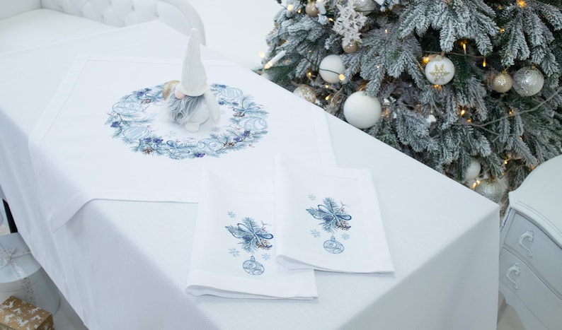 K&ouml;nnte beinhalten: Ein wei&szlig;es Tischtuch mit einem blau bestickten Weihnachtskranz und passenden Servietten. Eine Zwergfigur sitzt auf dem Tisch. Ein Weihnachtsbaum mit Ornamenten befindet sich im Hintergrund.