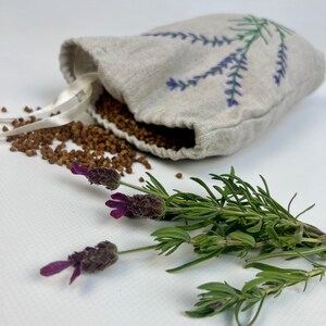 K&ouml;nnte beinhalten: Ein kleines, beiges Leinen-S&auml;ckchen, bestickt mit lila Lavendelzweigen, aus dem braune K&ouml;rner quellen. Frische Lavendelzweige mit lila Bl&uuml;ten und gr&uuml;nen Stielen sind in der N&auml;he angeordnet. Das S&auml;ckchen hat ein wei&szlig;es Band.