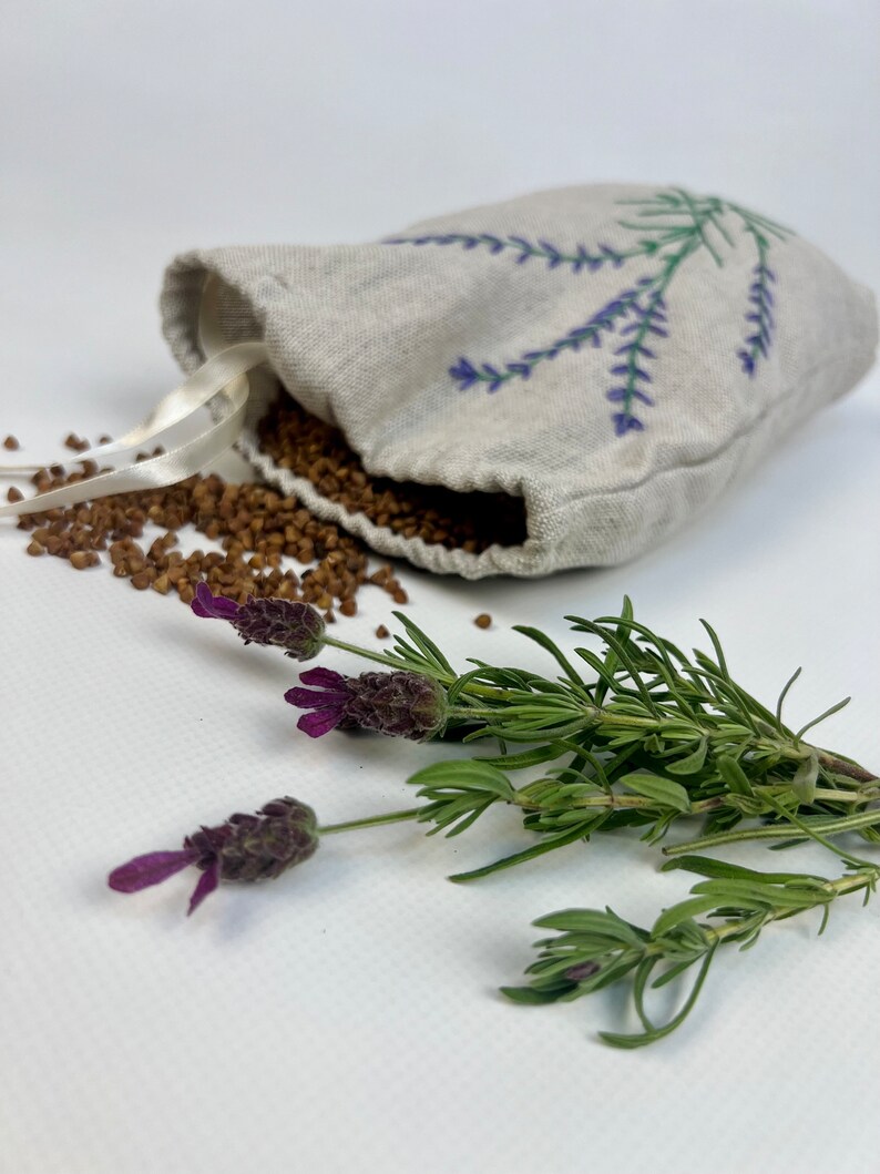 K&ouml;nnte beinhalten: Ein kleines, beiges Leinen-S&auml;ckchen, bestickt mit lila Lavendelzweigen, aus dem braune K&ouml;rner quellen. Frische Lavendelzweige mit lila Bl&uuml;ten und gr&uuml;nen Stielen sind in der N&auml;he angeordnet. Das S&auml;ckchen hat ein wei&szlig;es Band.
