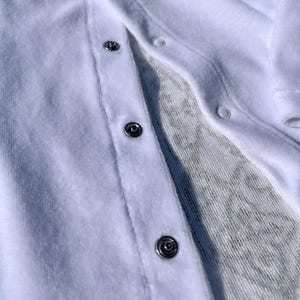 K&ouml;nnte beinhalten: Nahaufnahme eines wei&szlig;en Baby-Stramplers mit Druckkn&ouml;pfen. Der Strampler besteht aus einem weichen, strukturierten Stoff und hat mehrere silberne Druckkn&ouml;pfe auf der Vorderseite. Der Innenstoff hat ein dezentes, gemustertes Design.