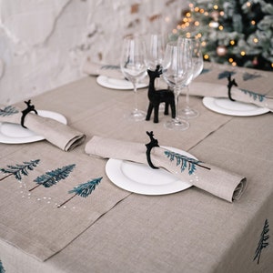 May include: A festive table setting with a beige linen tablecloth and matching runner. White plates hold rolled napkins secured by black reindeer napkin rings. Embroidered blue and white trees decorate the runner and napkins. Wine glasses and a black reindeer figurine complete the holiday scene.
