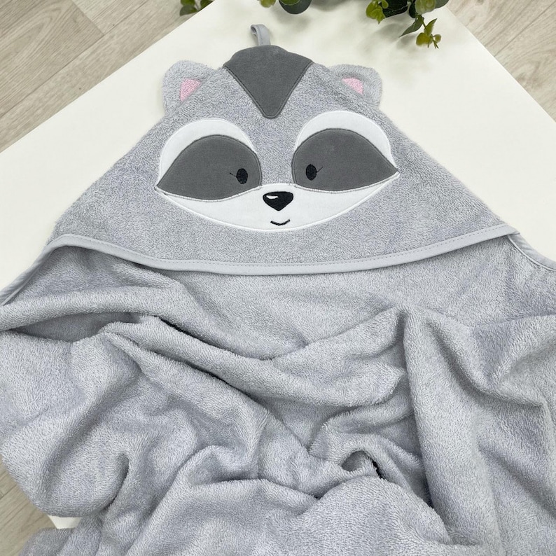 May include: A grey hooded baby towel featuring a raccoon face design. The hood has a raccoon's face with white, black, and pink details. The towel is made of soft, absorbent material for bath time.