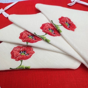 May include: Four white linen napkins with red embroidered poppies. The napkins are folded in a fan shape and are on a red background.