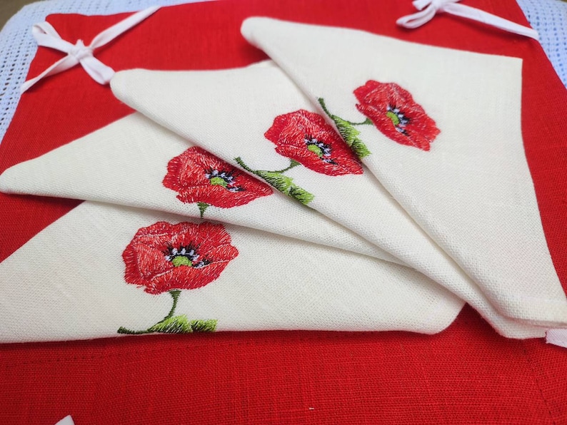 May include: Four white linen napkins with red embroidered poppies. The napkins are folded in a fan shape and are on a red background.