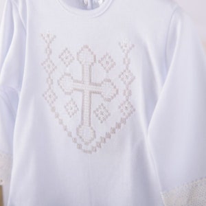 May include: White long-sleeved baby onesie with a white cross embroidered on the front. The cross is surrounded by a decorative pattern.