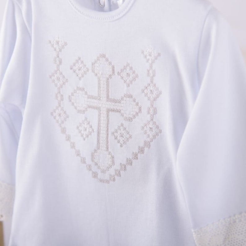 May include: White long-sleeved baby onesie with a white cross embroidered on the front. The cross is surrounded by a decorative pattern.