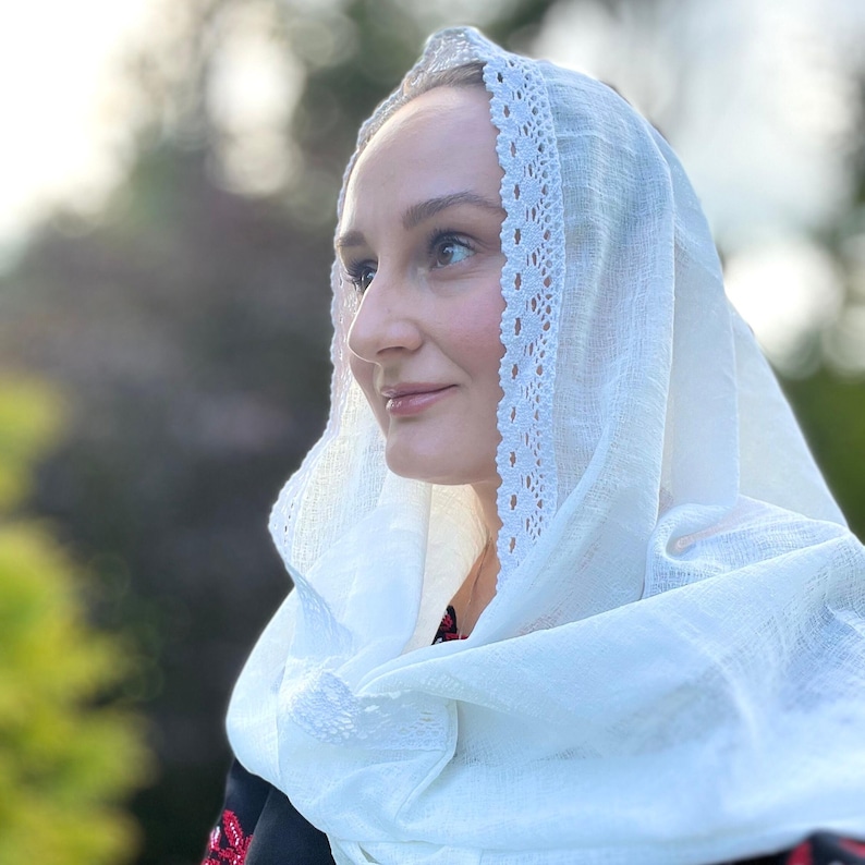 May include: A white headscarf with a delicate lace trim. The scarf is draped over the head and shoulders. The fabric appears to be lightweight and slightly sheer. The background is blurred, suggesting an outdoor setting.