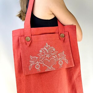 Op de afbeelding: Een koraalrode tote bag met een groot voorvak. Het voorvak heeft een wit geborduurd bloemenmotief met een hart in het midden.