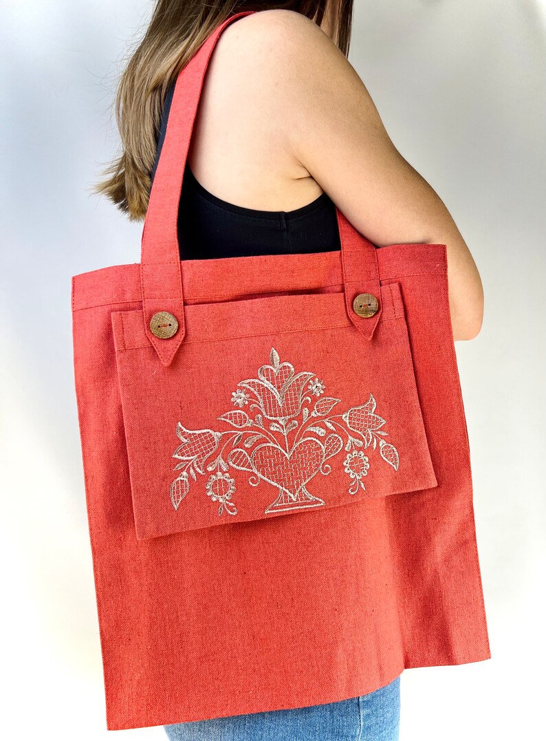 Op de afbeelding: Een koraalrode tote bag met een groot voorvak. Het voorvak heeft een wit geborduurd bloemenmotief met een hart in het midden.
