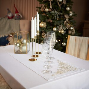 May include: A festive table setting with a white tablecloth, gold candle holders with white candles, and wine glasses. A gold-trimmed Christmas tree runner and a glass box with ornaments add to the holiday decor.