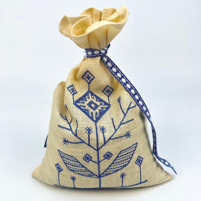 Pode incluir: Uma bolsa de linho bege com um desenho floral bordado em azul. A bolsa &eacute; amarrada com uma fita azul e branca. A parte superior da bolsa &eacute; franzida e amarrada com tecido bege.
