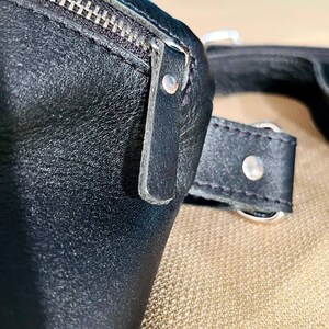 May include: Close-up of a black leather bag with a zipper and a strap. The bag has a silver buckle and a silver rivet.