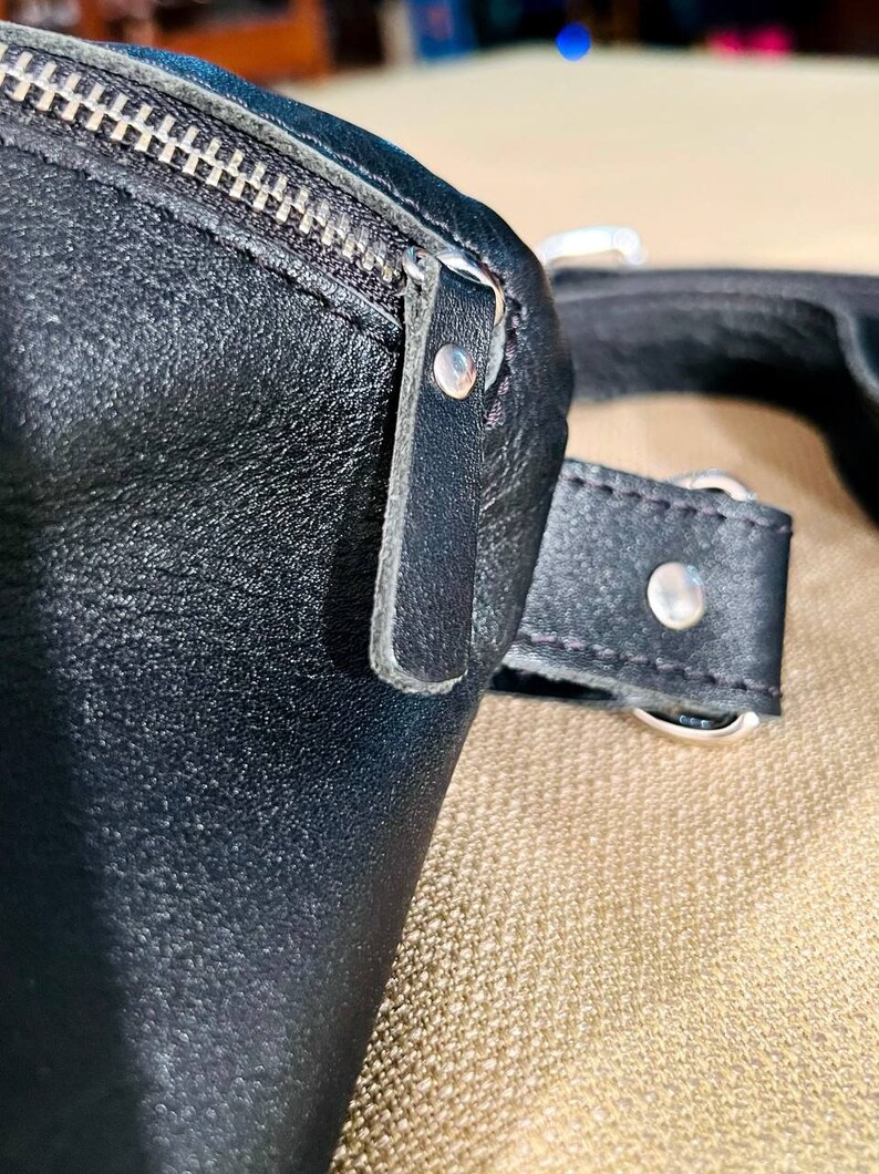 May include: Close-up of a black leather bag with a zipper and a strap. The bag has a silver buckle and a silver rivet.