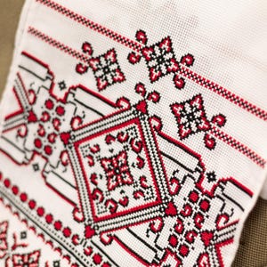 Puede incluir: Tela blanca con intrincado bordado de punto de cruz en rojo y negro. El dise&ntilde;o presenta patrones geom&eacute;tricos, motivos florales y bordes. El textil es probablemente una pieza decorativa o parte de ropa tradicional.