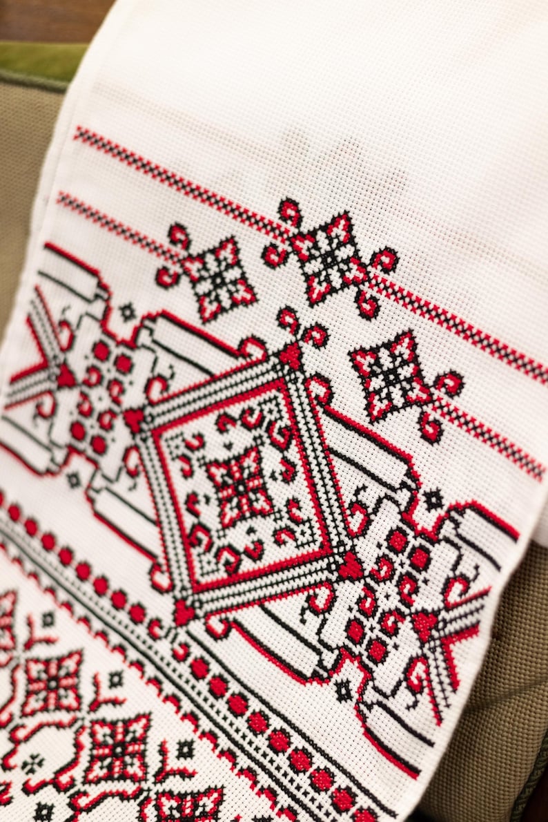 Puede incluir: Tela blanca con intrincado bordado de punto de cruz en rojo y negro. El dise&ntilde;o presenta patrones geom&eacute;tricos, motivos florales y bordes. El textil es probablemente una pieza decorativa o parte de ropa tradicional.
