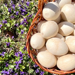 K&ouml;nnte beinhalten: Ein Weidenkorb gef&uuml;llt mit zahlreichen hellfarbigen, oval geformten Holzeiern. Der Korb ist braun und liegt zwischen gr&uuml;nem Laub und kleinen lila Blumen. Die Eier haben eine glatte, nat&uuml;rliche Oberfl&auml;che.