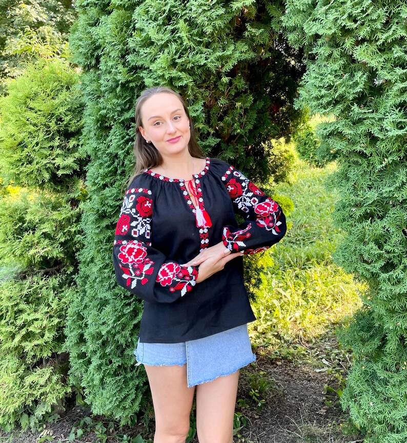 Puede incluir: Una blusa negra bordada con motivos florales rojos y blancos. La blusa tiene un escote redondo y mangas largas. La modelo lleva una falda vaquera.