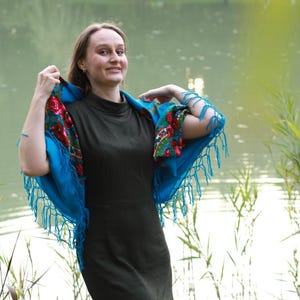 May include: A woman wearing a dark green dress and a bright blue shawl with floral patterns and fringe. The shawl is draped over the shoulders, creating a striking contrast against the dress. The background features a lake and greenery.