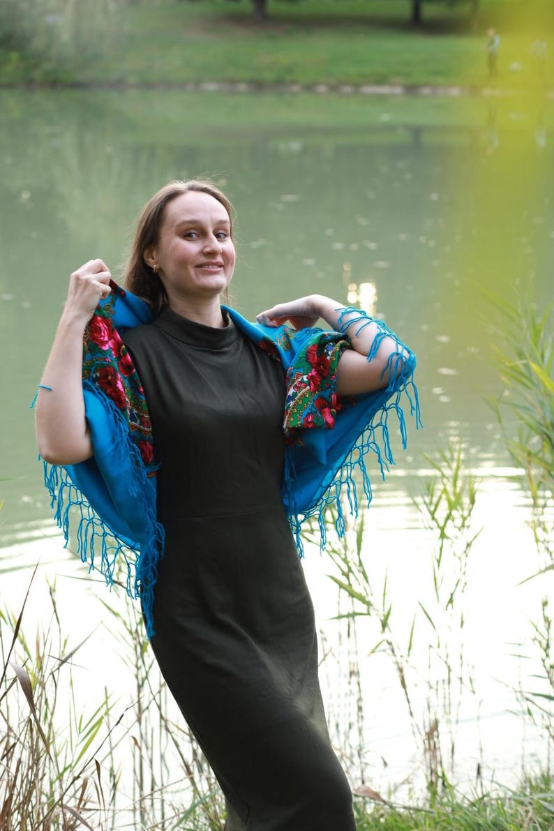 May include: A woman wearing a dark green dress and a bright blue shawl with floral patterns and fringe. The shawl is draped over the shoulders, creating a striking contrast against the dress. The background features a lake and greenery.