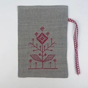 Puede incluir: Una bolsa gris de lino con cord&oacute;n y un dise&ntilde;o de &aacute;rbol bordado en borgo&ntilde;a. El &aacute;rbol tiene un patr&oacute;n geom&eacute;trico y est&aacute; centrado en la bolsa. Una cinta burdeos con lunares blancos est&aacute; sujeta en la parte superior.