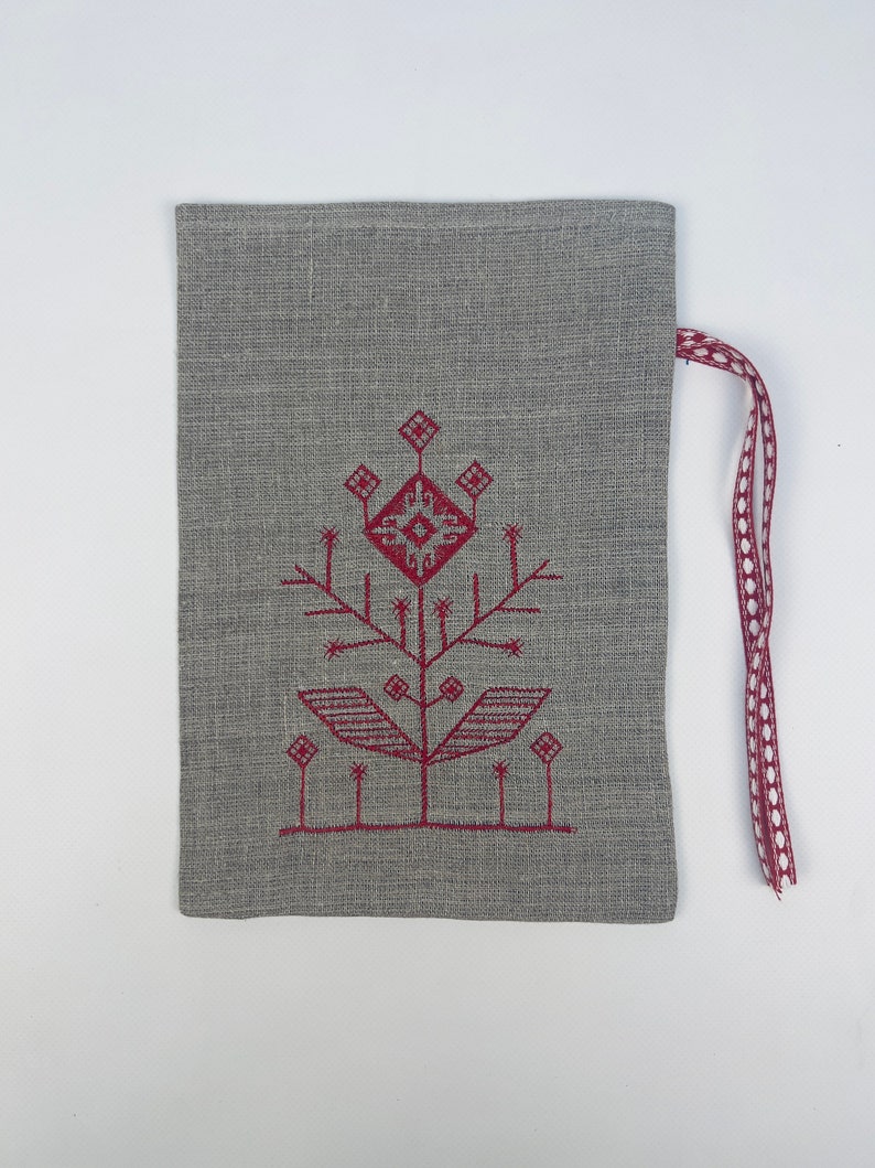 Puede incluir: Una bolsa gris de lino con cord&oacute;n y un dise&ntilde;o de &aacute;rbol bordado en borgo&ntilde;a. El &aacute;rbol tiene un patr&oacute;n geom&eacute;trico y est&aacute; centrado en la bolsa. Una cinta burdeos con lunares blancos est&aacute; sujeta en la parte superior.