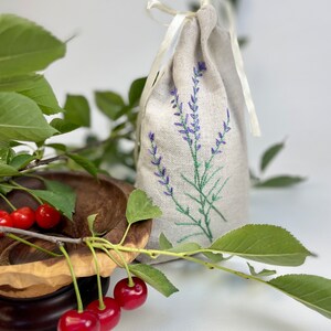 K&ouml;nnte beinhalten: Ein Leinens&auml;ckchen mit gestickten Lavendelzweigen in Lila, gebunden mit einem cremefarbenen Band. Frische rote Kirschen und gr&uuml;ne Bl&auml;tter sind neben einer Holzschale angeordnet. Das S&auml;ckchen ist mit Lavendel gef&uuml;llt.