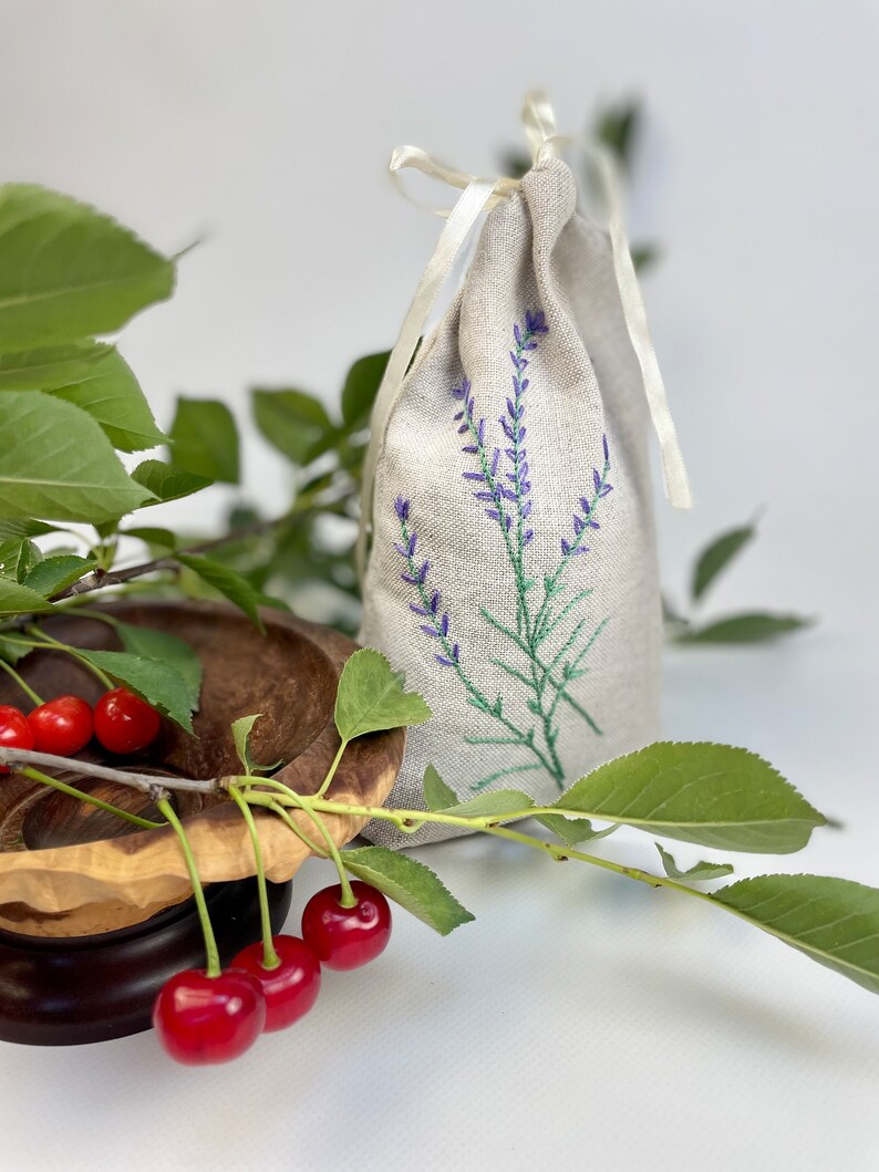 K&ouml;nnte beinhalten: Ein Leinens&auml;ckchen mit gestickten Lavendelzweigen in Lila, gebunden mit einem cremefarbenen Band. Frische rote Kirschen und gr&uuml;ne Bl&auml;tter sind neben einer Holzschale angeordnet. Das S&auml;ckchen ist mit Lavendel gef&uuml;llt.