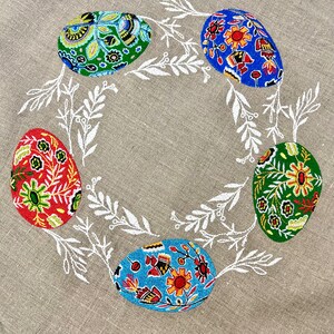 以下が含まれることがあります： ベージュのリネン製のテーブルクロスに、白い刺繍で葉とカラフルなイースターエッグ5個がデザインされています。卵は赤、青、緑、黄色の花柄で飾られています。