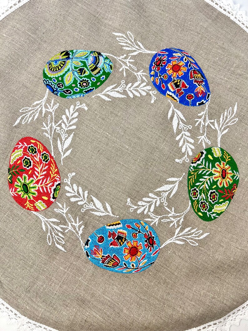 以下が含まれることがあります： ベージュのリネン製のテーブルクロスに、白い刺繍で葉とカラフルなイースターエッグ5個がデザインされています。卵は赤、青、緑、黄色の花柄で飾られています。
