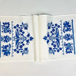 Peut inclure: Chemin de table en lin blanc avec un motif floral bleu d&eacute;taill&eacute;. Le design comprend des fleurs, des feuilles et des oiseaux stylis&eacute;s. Les bords sont finis avec une bordure crochet&eacute;e bleu fonc&eacute;, ajoutant une touche d&eacute;corative.