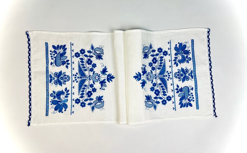 Peut inclure: Chemin de table en lin blanc avec un motif floral bleu d&eacute;taill&eacute;. Le design comprend des fleurs, des feuilles et des oiseaux stylis&eacute;s. Les bords sont finis avec une bordure crochet&eacute;e bleu fonc&eacute;, ajoutant une touche d&eacute;corative.