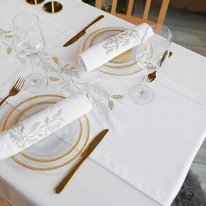 Puede incluir: Un mantel blanco con dise&ntilde;os florales bordados, con cubiertos dorados, copas de vino transparentes y servilletas enrolladas. La mesa est&aacute; puesta con platos transparentes con bordes dorados, creando un arreglo elegante.