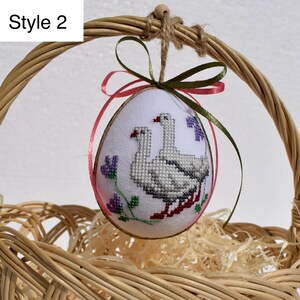 Op de afbeelding: Een geborduurd paasei met een kruissteekontwerp van twee ganzen, paarse bloemen en rode accenten. Het ei wordt opgehangen met een groen en roze lint en bevindt zich in een rieten mand. De tekst "Style 2" staat linksboven.