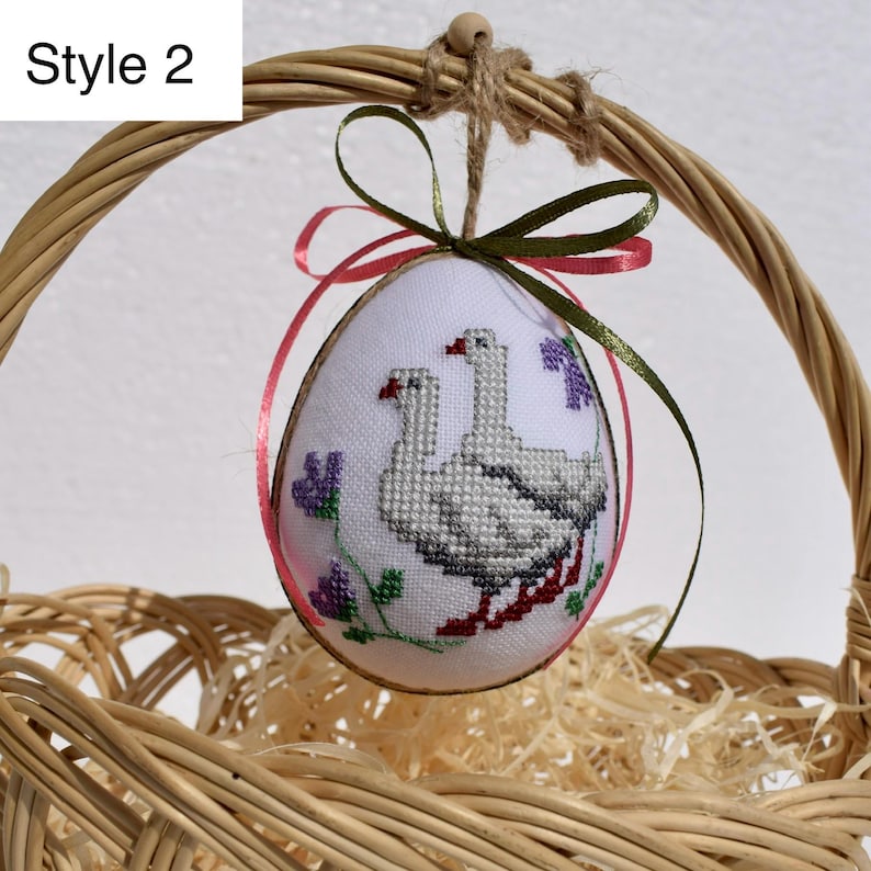 Op de afbeelding: Een geborduurd paasei met een kruissteekontwerp van twee ganzen, paarse bloemen en rode accenten. Het ei wordt opgehangen met een groen en roze lint en bevindt zich in een rieten mand. De tekst "Style 2" staat linksboven.