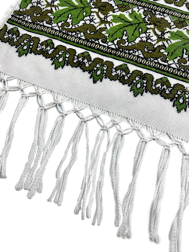 May include: White textile with a green and olive leaf pattern and a fringe. The leaf design is repeated across the top of the textile. The fringe is made of white threads.