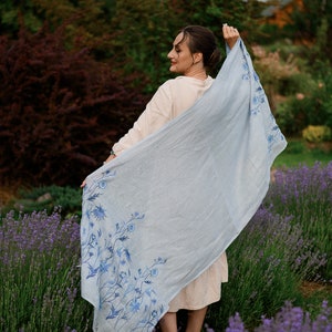 K&ouml;nnte beinhalten: Eine Frau in einem hellbeigen Kleid h&auml;lt einen hellblauen Schal mit Blumenmuster. Der Schal hat ein blaues Blumenmuster am unteren Rand. Die Frau steht auf einem Lavendelfeld.
